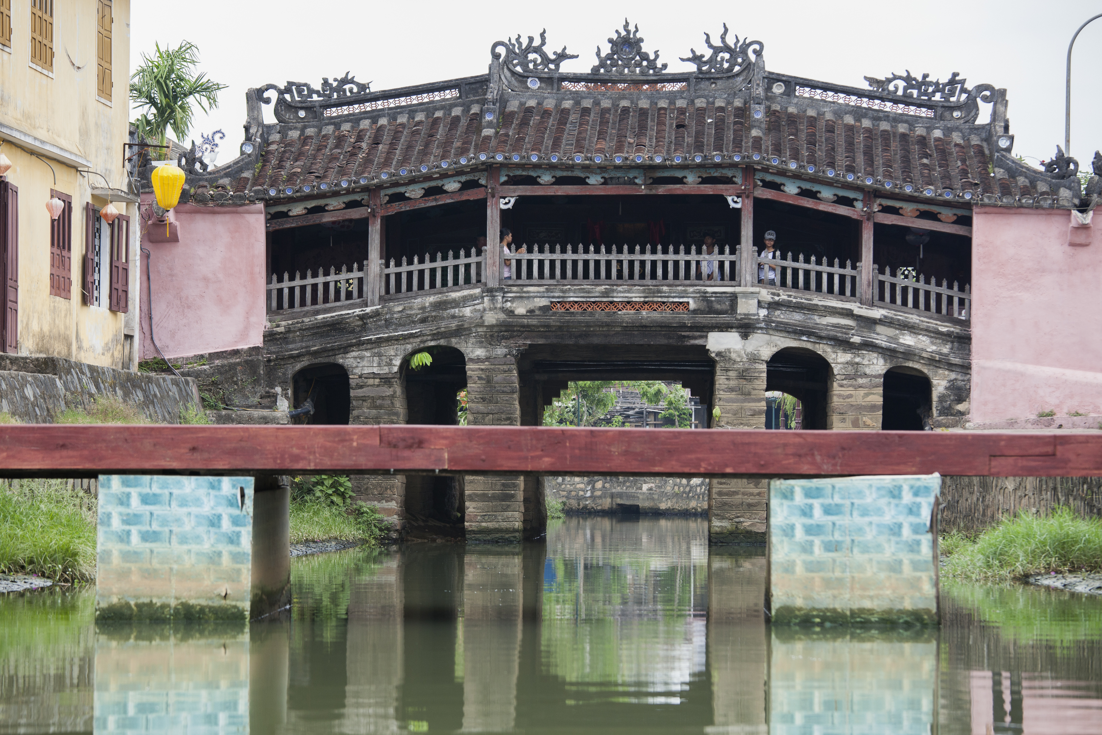 The iconic Japanese Covered Bridge in Hoi An, with its distinctive red-orange arched roof, crossing a small canal under a clear blue sky.