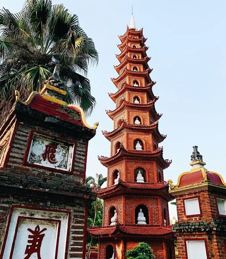 The Tran Quoc Pagoda, an ancient Buddhist temple with multiple red-tiered stupas, situated on an islet in Hanoi's West Lake at sunset, with reflections on the water.