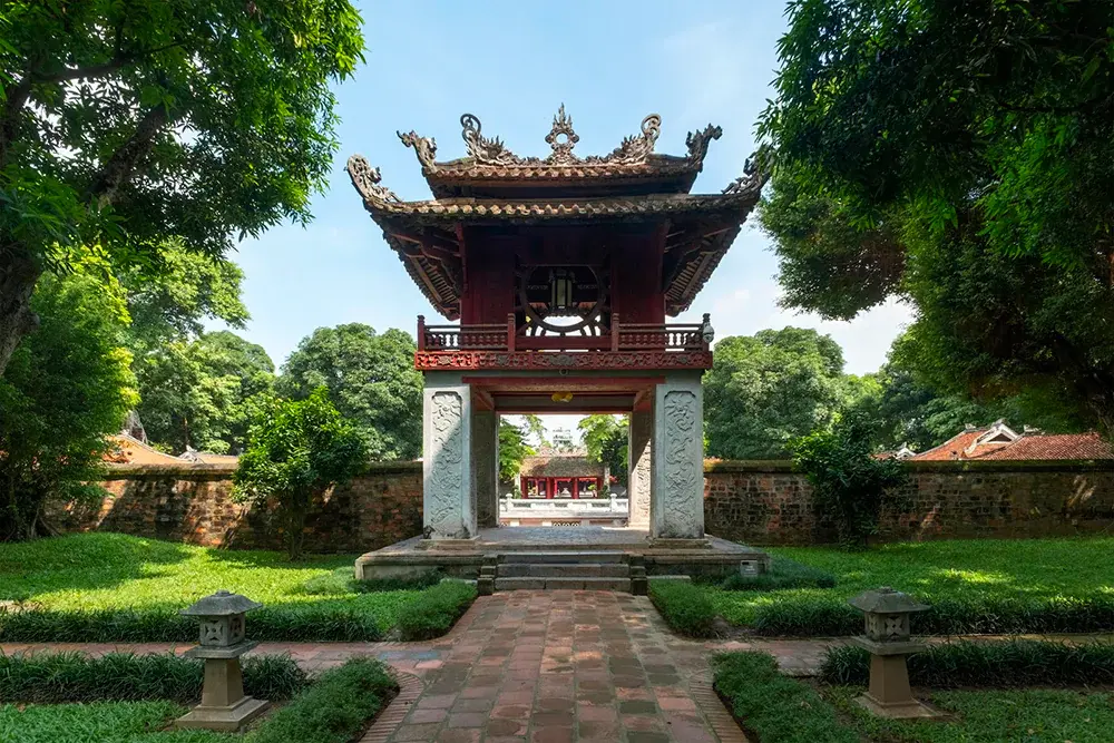 The Temple of Literature, a traditional Vietnamese architectural complex with red gates, courtyards, and ancient trees, under a clear sky.